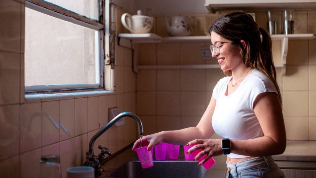 A young woman in glasses washes dishes by a window in a vintage-style kitchen.